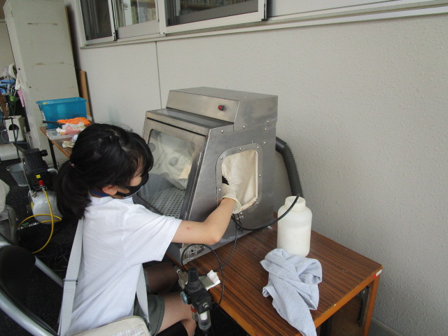 屋外の軒下で、白いTシャツを着てマスクを着用した女の子が、手袋を介して金属製の箱型装置に手を入れて中の物を操作している作業風景の写真