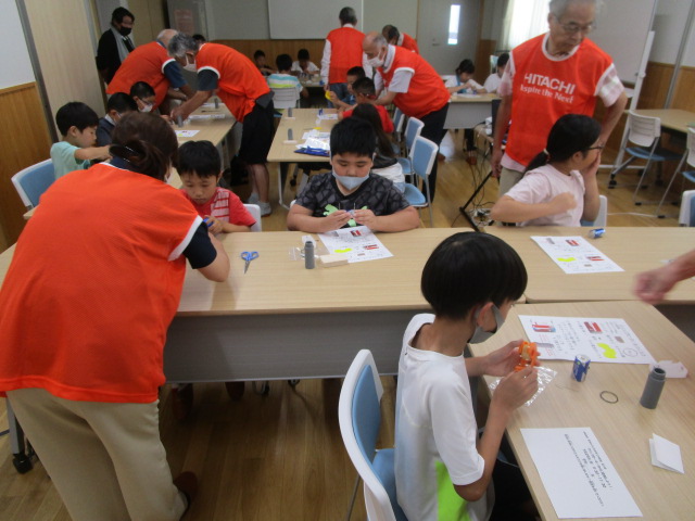 オレンジ色のベストを着用した複数の大人たちが、室内で小学生たちに工作の指導を行い、子どもたちが机に向かい小さな部品を組み立てている活気あるワークショップ風景の写真