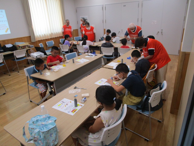 広い室内で、オレンジ色のベストを着用した複数の大人たちが、小学生の子どもたちにリニアモーターカーの簡易模型作りを指導し、子どもたちが熱心に工作に取り組んでいるワークショップ風景の写真