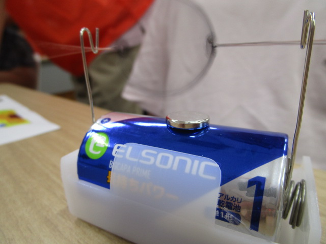 机の上で、単一電池に磁石を取り付け、その両端にコイル状のバネと針金を接続してモーターの仕組みを簡易的に再現している実験装置をクローズアップした写真