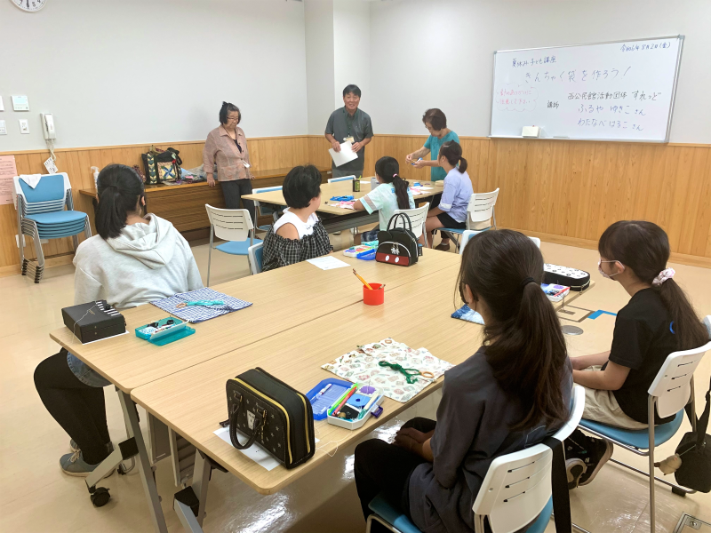 「夏休み手芸講座 きんちゃく袋を作ろう！」というテーマのボードがかけられた室内で、数名の大人と子どもたちがテーブルを囲んで手芸に取り組むワークショップの様子の写真