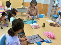テーブルを囲んで座る複数の子どもたちが、和柄や花柄などの布を使って裁縫に取り組んでおり、中央では女性が優しく手芸の指導をしている様子の写真