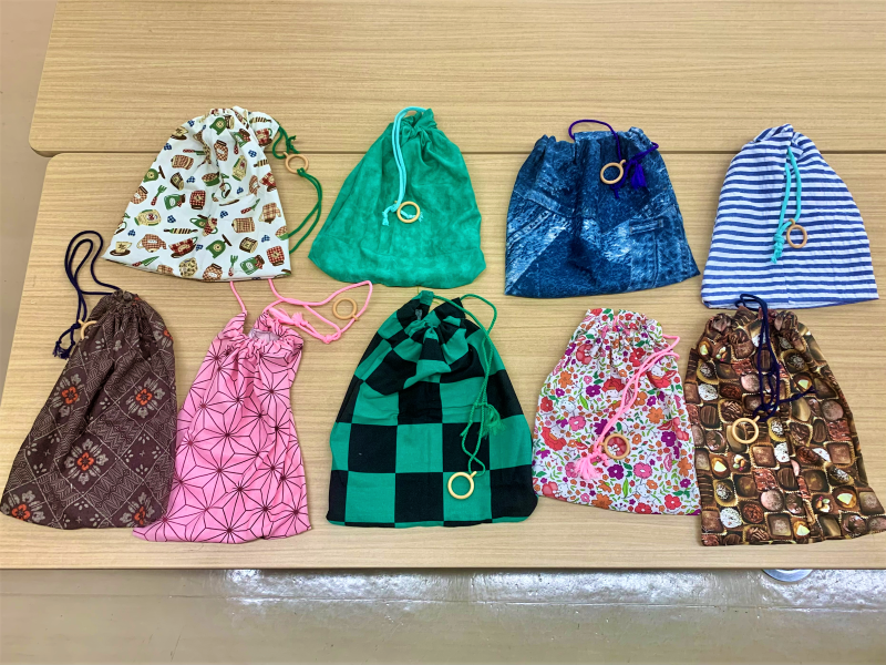 木目調のテーブルの上に、茶器柄、和柄、市松模様、花柄など異なる柄の布地で手作りされた巾着袋が、上部にひもと木製リングを付けたられ並べられている集合写真