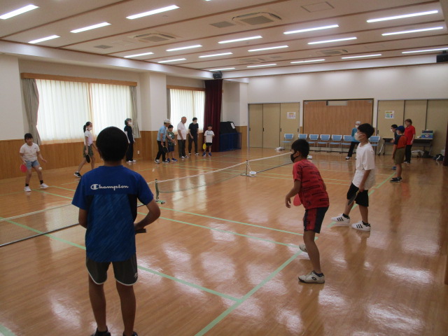 木製のフローリングが敷かれた広い室内で、ネットを挟んで向かい合い、子どもたちが大人とテニスを楽しんでいるスポーツ活動の様子の写真