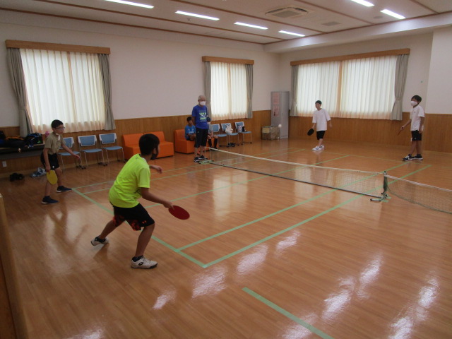緑色のラインを引いたフローリングの体育館のような室内で、4人の子どもたちがラケットを持ってネット越しに向かい合いフリーテニスを楽しんでいる写真