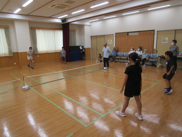 明るいフローリングの広い室内で、ネットを設置してフリーテニスを楽しむ子どもたちを、数名の高齢者と大人が見守っているスポーツ交流風景の写真