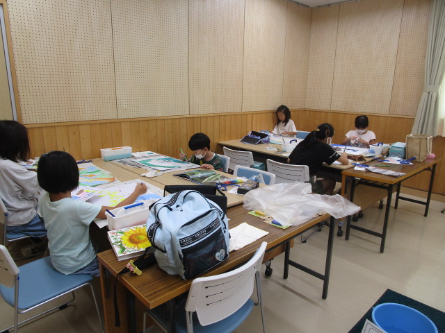 会議室のような木目と白の壁で構成された室内で、複数のテーブルに分かれて数人の子どもたちが、絵の具や色鉛筆を使って集中して絵画や工作に取り組むワークショップ風景の写真