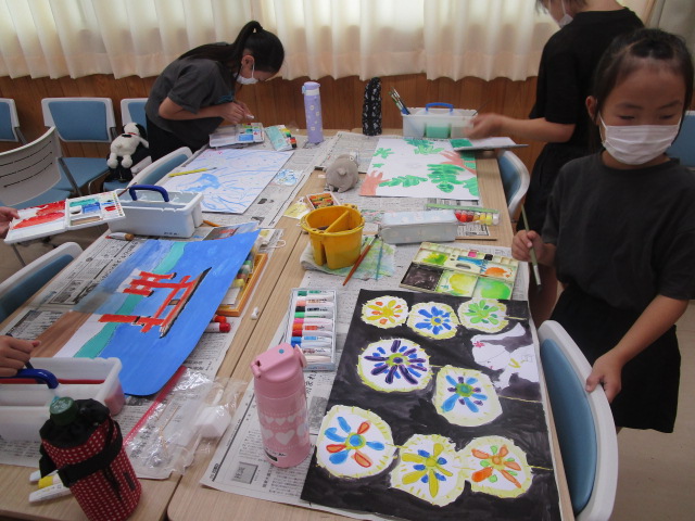室内でマスクを着用した子どもたちが、新聞紙を敷いた机の上で水彩絵の具やパステルを使い、鳥居や花火など様々な題材の絵画制作に熱心に取り組むアート教室の様子の写真