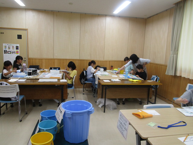 薄いベージュと木目調の壁の室内で、複数のテーブルに分かれて子どもたちが工作や絵画などの創作活動に取り組み、大人ひとりが指導をしている様子の写真