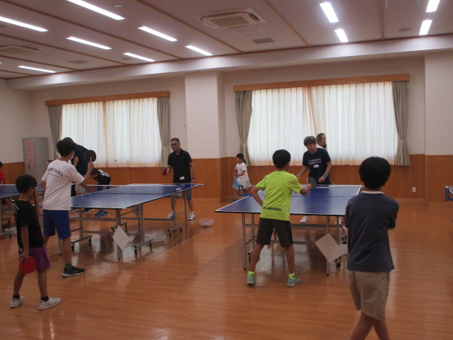 複数の卓球台を囲み、子どもたちが大人と共に卓球を楽しんでいるスポーツ教室の様子の写真