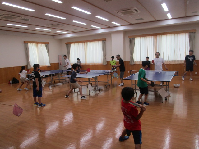 光沢のあるフローリングの室内で、複数の卓球台を使って、子どもたちが数名の大人と共に卓球の練習に取り組んでいる活動風景の写真