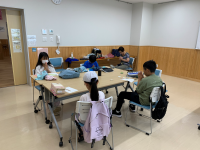 会議室のような明るい室内で、木目調のテーブルを囲んで4人の児童たちが学習や作業に静かに取り組んでいる様子の写真