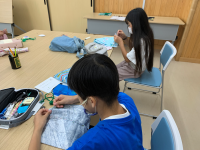 白いマスクを着用した2人の子どもが、木目調のテーブルで裁縫道具や布を使って、真剣な表情で何かの工作や手芸に取り組んでいる作業風景を捉えた写真