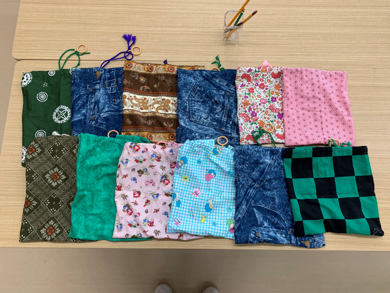 テーブルの上に、和柄、デニム調、花柄、キャラクター柄など多彩な生地で作成された巾着袋が並べられている集合写真