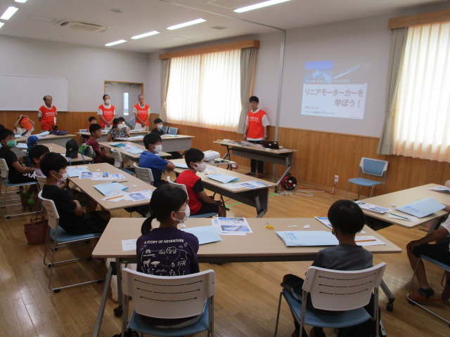 教室で複数人の講師と多数の小中学生が参加し、プロジェクターに「リニアモーターカーを学ぼう！」というタイトルが映し出されている学習会を開催している様子の写真