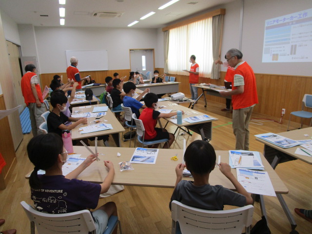 オレンジ色のベストを着た大人が複数名、子どもたちにリニアモーターカーの工作指導を行っているワークショップ風景を捉えた写真