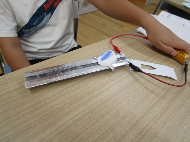 Tシャツ姿の子どもが、アルミホイルを貼ったレールと単三電池をつなぎ、クリップ付きコードと磁石を用いてリニアモーターカーの簡易模型を自作している手元を写した写真