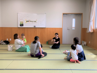 畳敷きの明るい部屋で、母親と子どもたちが輪になって座り、黒い服を着た女性が話している様子を捉えた写真