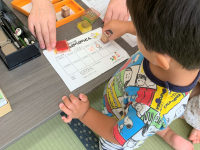 幼児が、大人の手助けを受けながらテーブルの上にある出席確認のシートにスタンプを押している様子を上方から捉えた写真