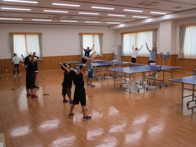 手前の子供たちや奥の大人たちが、両腕を頭上で組んで体を左右に倒すストレッチ運動を全員で熱心に行っている準備運動の様子の写真