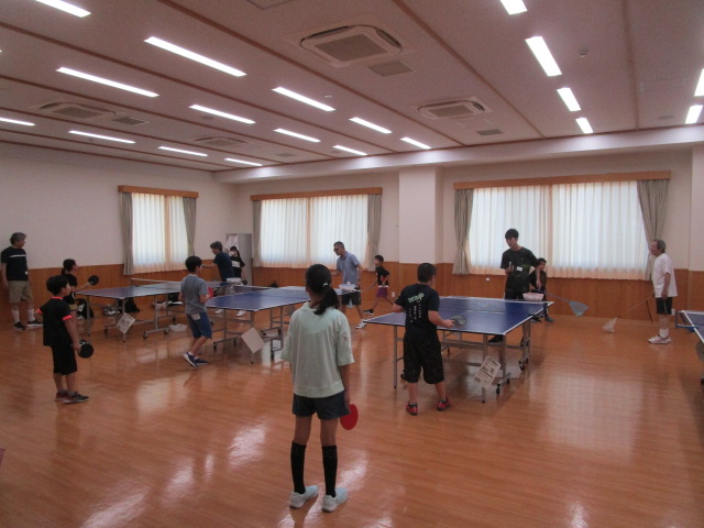 フローリングの広い部屋に複数の卓球台が設置され、子供たちと大人がそれぞれの台で卓球のラリーや指導を受けており、全員が熱心に活動に取り組んでいる、卓球教室の活気ある練習風景を捉えた写真