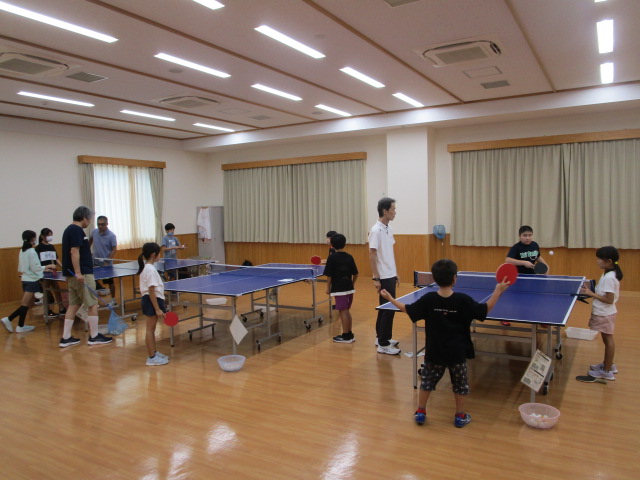 多くの卓球台が並ぶフローリングの広い部屋で、子供たちと大人がそれぞれの台で卓球の練習や指導を受けており、全員が熱心に活動に取り組んでいる、卓球教室の活気ある練習風景を捉えた写真