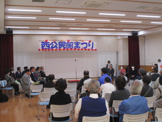 明るい室内ホールで、正面の白い壁に「西公民館まつり」という青と赤の横断幕が掲げられ、手前に複数の参加者が白い椅子に座り、中央前方に数名の登壇者が立って挨拶を行っている公民館イベントの開会式の写真