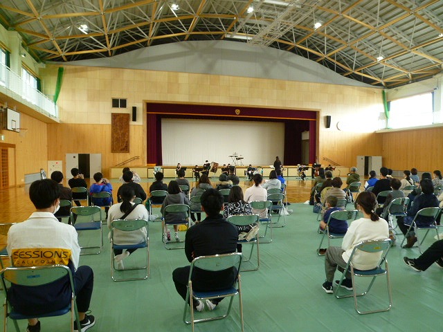 高い天井に金属トラスと照明が並ぶ体育館で、緑色の床に白い椅子が配置され、観客が着席して正面のステージに向かい、管楽器アンサンブルの演奏を鑑賞している写真