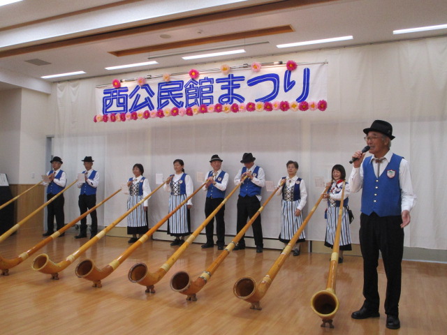 「西公民館まつり」という青と赤の横断幕の前に、黒い帽子と青いベスト、ストライプのエプロンを着た複数の演奏者が長いアルプホルンを床に立てて演奏し、右端の男性がマイクを持って話している民族楽器演奏会の写真