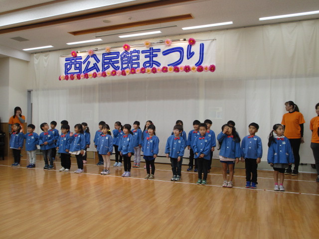 木製床の明るいホールで、「西公民館まつり」という青と赤の横断幕の前に、青い体操服を着た多数の子どもたちが整列して立ち、両端にオレンジ色のシャツを着た指導者が付き添っている発表会の写真