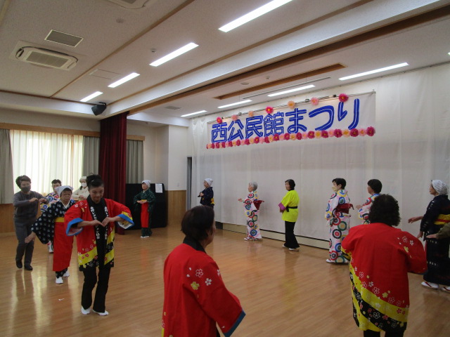 木製床の明るいホールで、「西公民館まつり」という青と赤の横断幕の前で、赤や黄色の鮮やかな衣装を着た複数の参加者がよさこい踊りを披露し、観客が左側と後方に立って観覧している写真