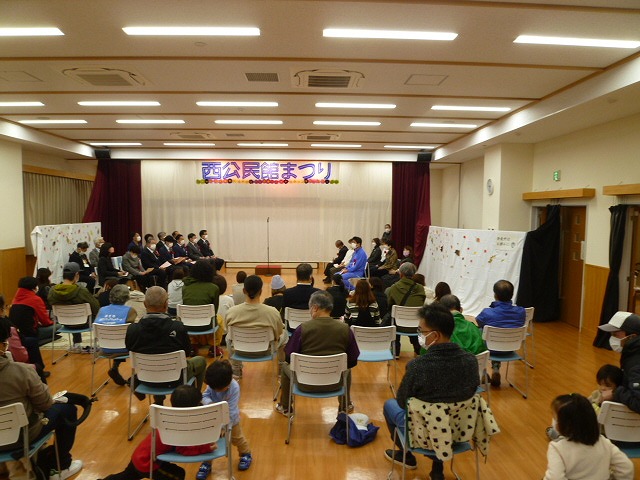 木製床のホールの正面ステージに「西公民館まつり」という紫色の横断幕が掲げられ、黒い着物姿の演奏者たちが琴を演奏し、手前に白い椅子に座った多数の観客が鑑賞している写真