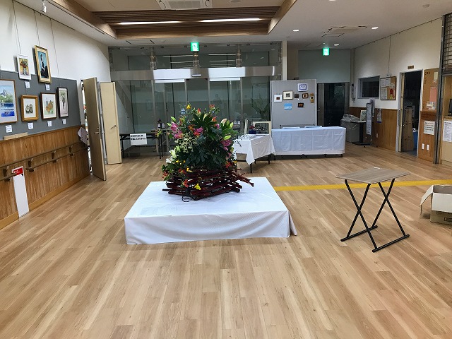 木製フローリングの明るいギャラリースペースで、中央に白い布がかけられたテーブルの上に華やかな生け花が飾られ、左側の壁に複数の額装作品が展示され、奥に白いテーブルクロスがかけられた受付台がある文化展示会場の写真