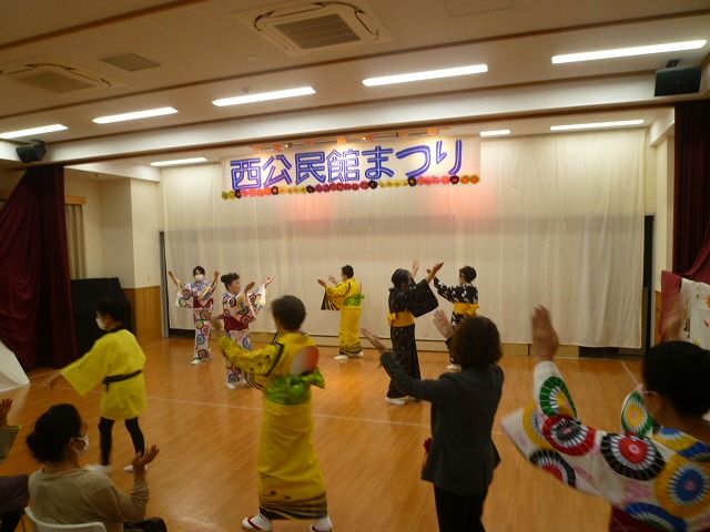 木製床のホールで、「西公民館まつり」という青と赤の横断幕の前で、黄色や花柄の浴衣を着た複数の参加者が扇子や鳴子を持って盆踊りを踊り、観客が手拍子で参加している写真
