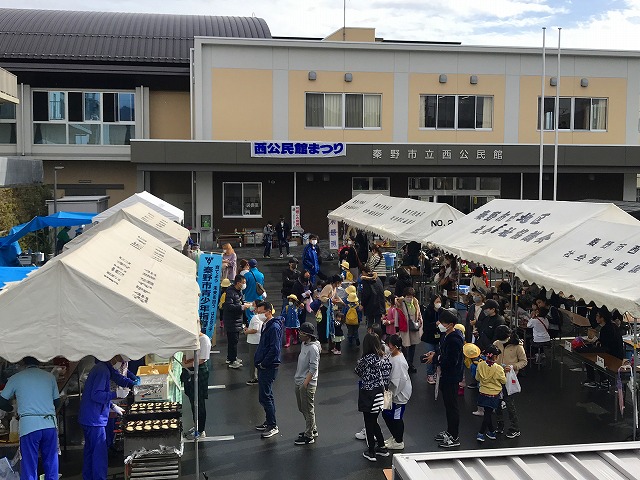 青空の下、「秦野市立西公民館」という看板のある薄茶色の建物前の広場に、白いテントが複数設置され、多数の来場者が黄色い帽子をかぶった子どもたちを含めて集まり、屋外イベントで飲食やゲームを楽しんでいる地域祭りの賑やかな風景写真