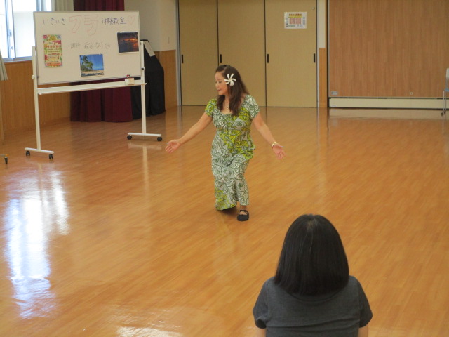 広いフローリングの部屋で、ハワイアンな花柄のドレスを着用し頭に白い花を飾った女性が優雅なポーズで立っており、手前には黒い服を着た生徒の後ろ姿が写っている指導の様子の写真