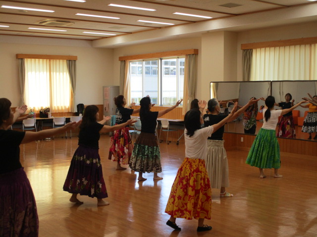 窓から光が差し込む明るいフローリングの広間の中で、カラフルなロングスカートを履いた女性たちが、鏡に向かって両手を広げるフラダンスの優雅なポーズをとって練習に励んでいる写真