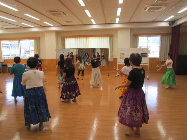 フローリングの広間の中で、カラフルなロングスカートを履いた女性たちが、講師に合わせて鏡の前に並んでフラダンスを踊っている、活気あふれるダンスレッスンの風景を捉えた写真