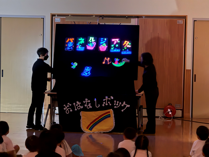 真っ暗なステージ上で、黒い服を着た二人の演者が、蛍光色の光る絵や文字が浮かび上がった黒いパネルを使って人形劇のようなものを演じており、床に座った大勢の子供たちがその様子を熱心に見つめている、おはなしポッケの公演風景の写真