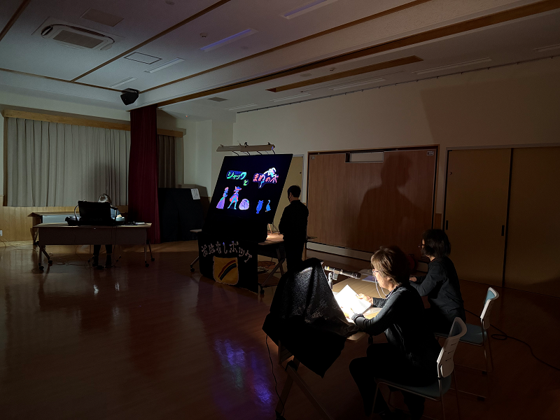 照明が落とされたフローリングの広間の中央で、黒い服の二人がパネルシアターのようなものを操作して光る人形を見せており、手前の机では二人の女性が作業をしている、おはなしポッケの上演中の静かな風景を捉えた写真