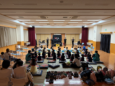 畳とフローリングの広間全体を写しており、中央の黒いパネルを使って行われる「おはなしポッケ」の公演を、大勢の子供たちが座布団の上に座って鑑賞し、その後ろで大人が見守っている賑やかな様子の写真