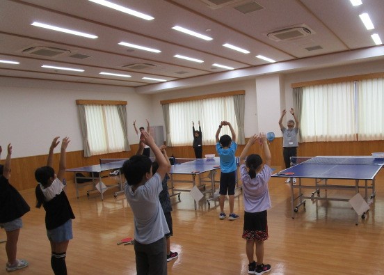 卓球台が複数置かれたフローリングの広い部屋で、手前の子供たち数名と奥の大人数名が、両手を上げて体を横に倒すストレッチ運動を全員で行っている、準備運動の様子の写真