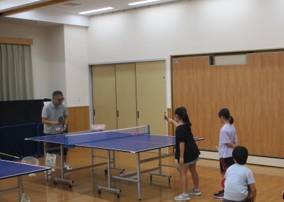 フローリングの広間の中央に置かれた卓球台を挟んで、灰色のシャツを着た男性と女の子が卓球の練習をしており、右奥には男の子の姿も見られる、卓球教室の和やかな様子の写真