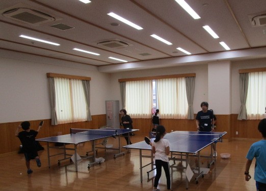 窓際の明るい部屋で、数台の卓球台が設置されており、黒い服を着た子供と白い服の女の子がそれぞれ卓球のラリーをしており、奥には数人の大人が立って練習の様子を見守っている、卓球の練習風景を捉えた写真