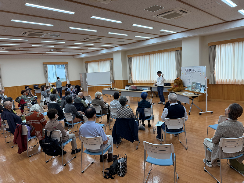 公民館のような広いフローリングの部屋で、高齢者を中心に大勢の聴衆が椅子に座って正面の講師の話を聞いており、ホワイトボードや大きなぬいぐるみが置かれた机が演台として使われている、講演会の様子の写真