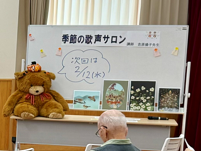 「季節の歌声サロン」と書かれたホワイトボードの前に、麦わら帽子を被った大きなテディベアのぬいぐるみと、数枚の風景や花の絵が飾られており、青いマジックで書かれた告知を、客席側から捉えた写真