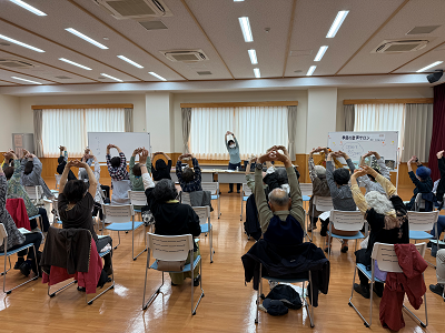 大勢の参加者が椅子に座り、両手を組んで頭上に大きく伸ばすストレッチ運動を、正面に立つ指導者と一緒に行っており、広々としたフローリングの部屋で和気藹々と健康づくりに励んでいる様子の写真