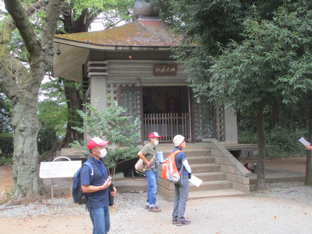 薄茶色の六角形の屋根を持つ、木造の小さな建物の前に、マスクを着用した3人の登山客のような人たちが立ち止まっており、周囲は背の高い木々に囲まれた自然豊かな場所を捉えた写真