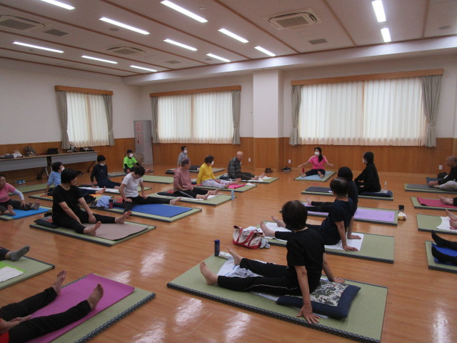 明るいフローリングの広間の中で、多くの参加者がヨガマットや座布団の上にあぐらをかいたり足を伸ばしたりして座っており、正面のピンクのシャツを着た指導者の動きを見ながら体操に励んでいる様子の写真