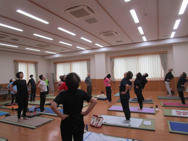 フローリングの広い部屋で、多数の参加者がヨガマットや座布団の上で立ち、腰に手を当てて上を見上げるポーズを取っており、全員が健康のために熱心に運動している様子の写真